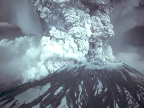 A towering grey column of ash rises from Mount St. Helens into a blue sky.